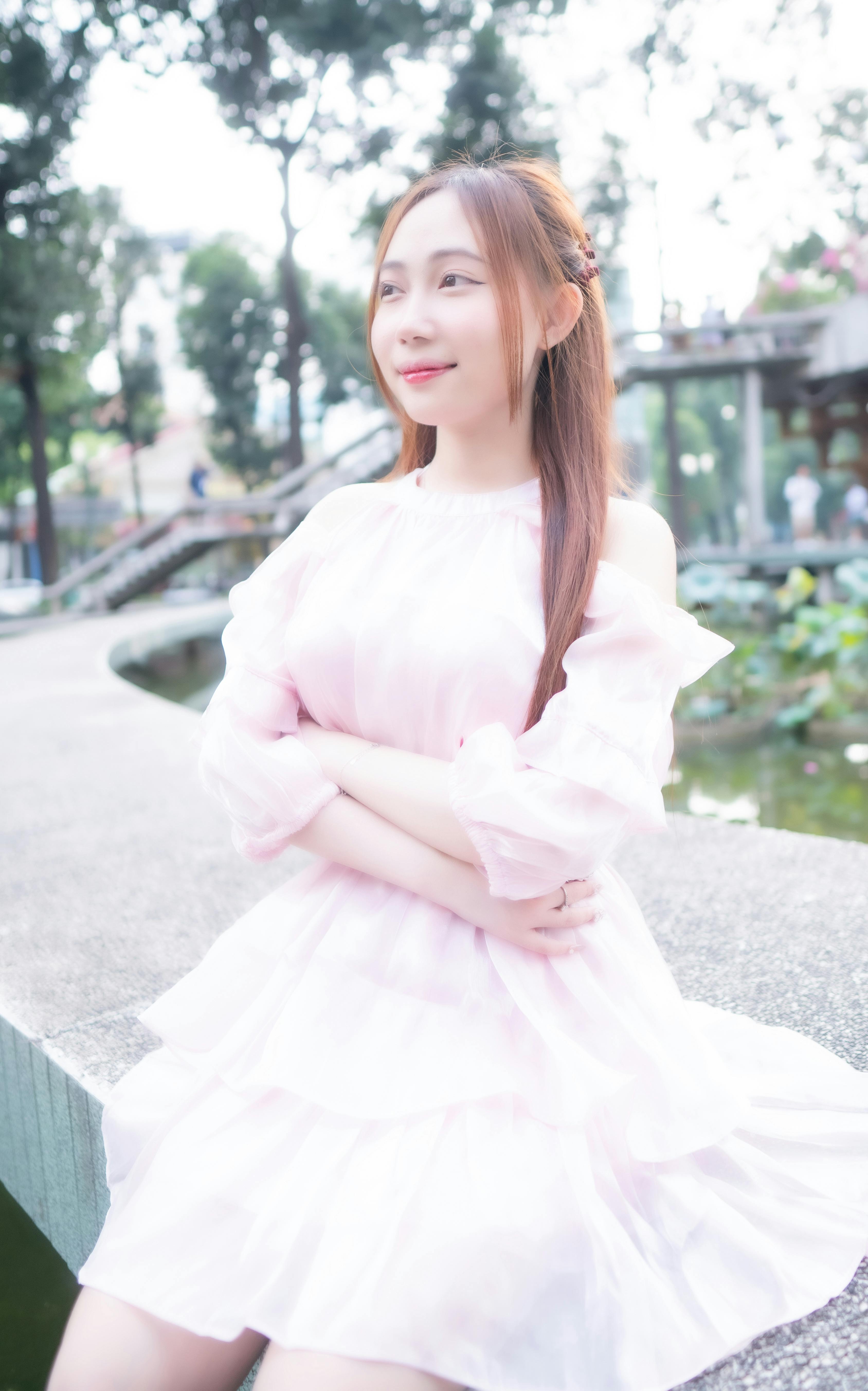 Young woman in a soft pink dress sitting by a serene pond in a lush park setting.