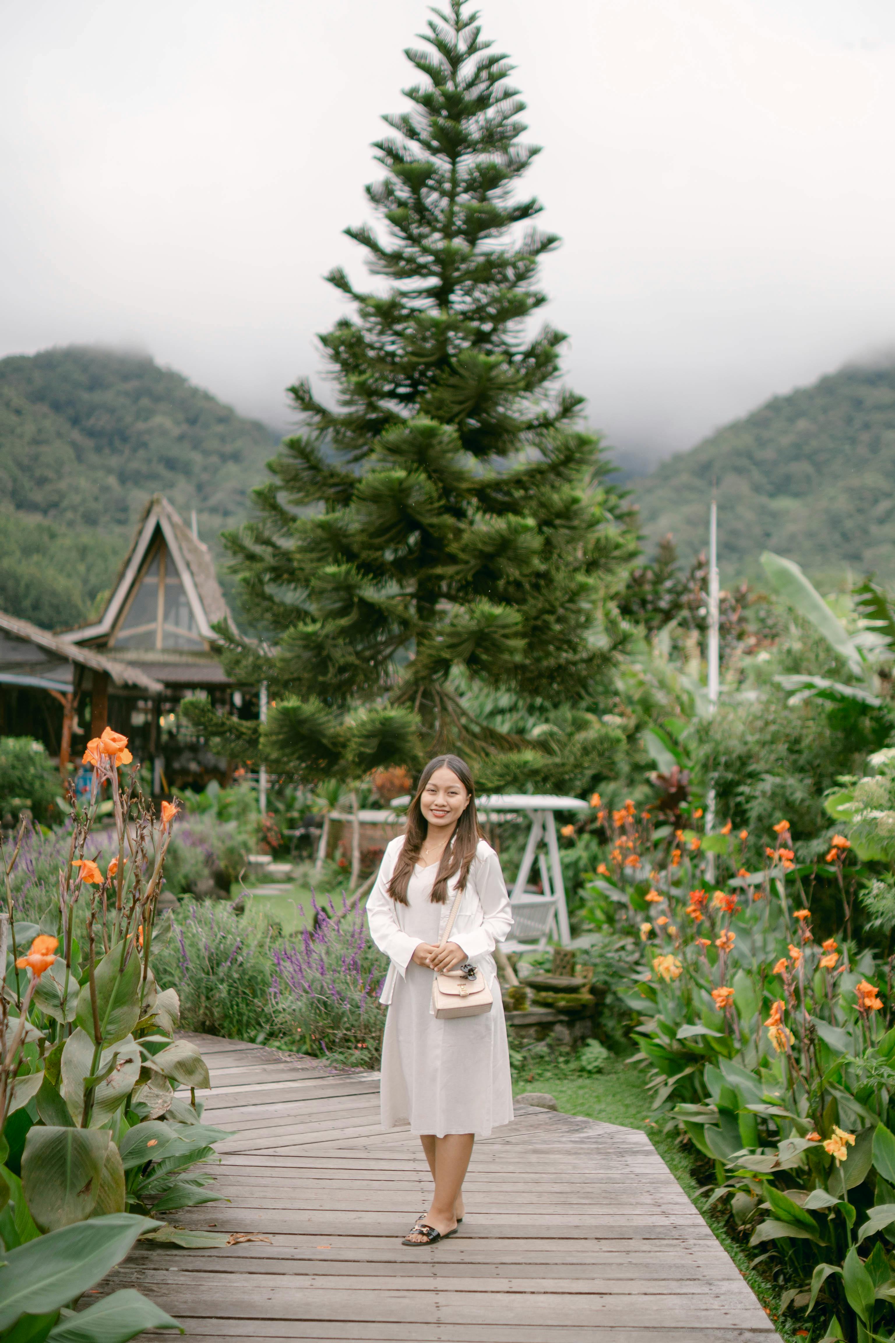 A young woman standing in a vibrant garden surrounded by flowers and greenery on a cloudy day.