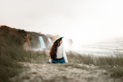 Free A young girl wearing a hat sits on a grassy sandy beach near a coastal waterfall. Stock Photo