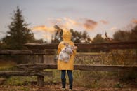 日落时分，一个孩子穿着黄色雨衣，拿着毛绒玩具站在乡村栅栏旁