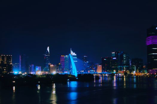 A dazzling night view of the modern skyline of Ho Chi Minh City, Vietnam with bright neon lights reflecting on water.