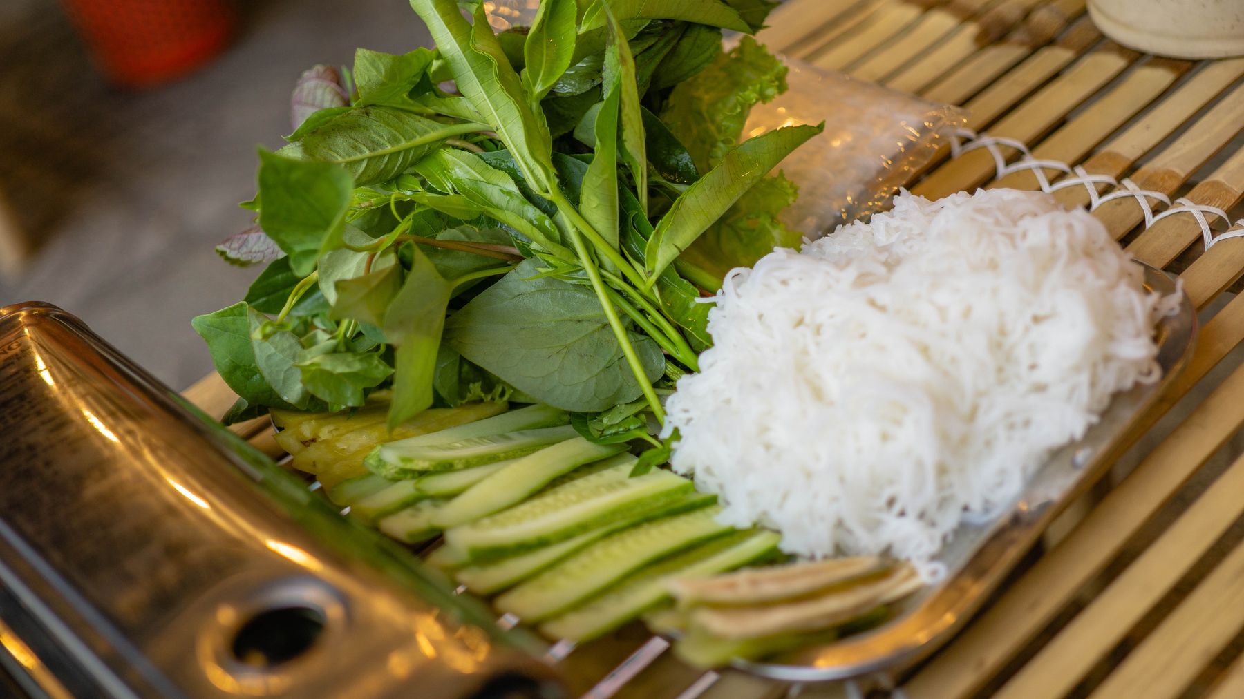 Herbs, rice noodles, and cucumbers on a bamboo table for a Vietnamese meal prep.