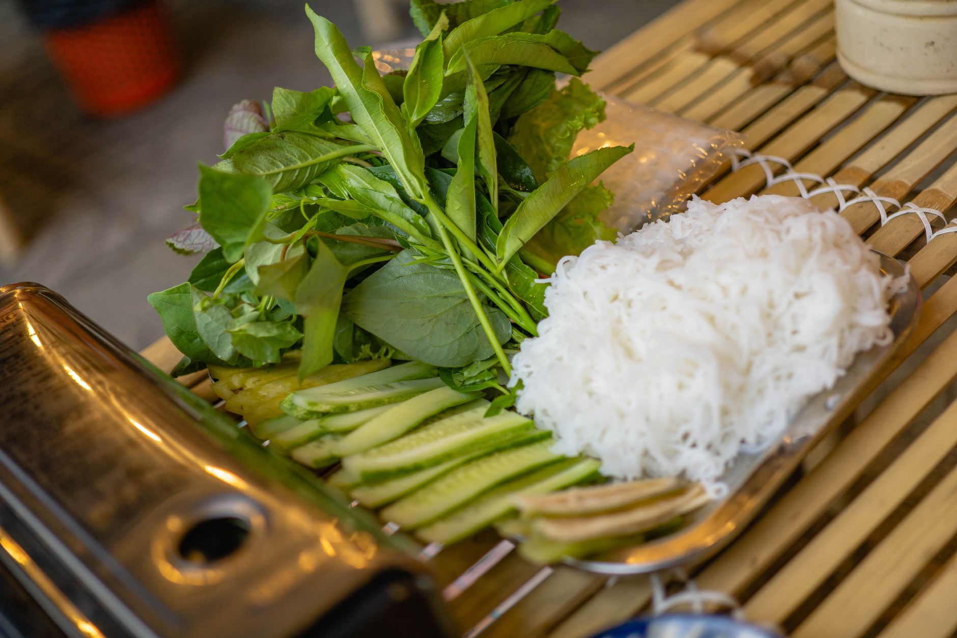 Herbs, rice noodles, and cucumbers on a bamboo table for a Vietnamese meal prep.