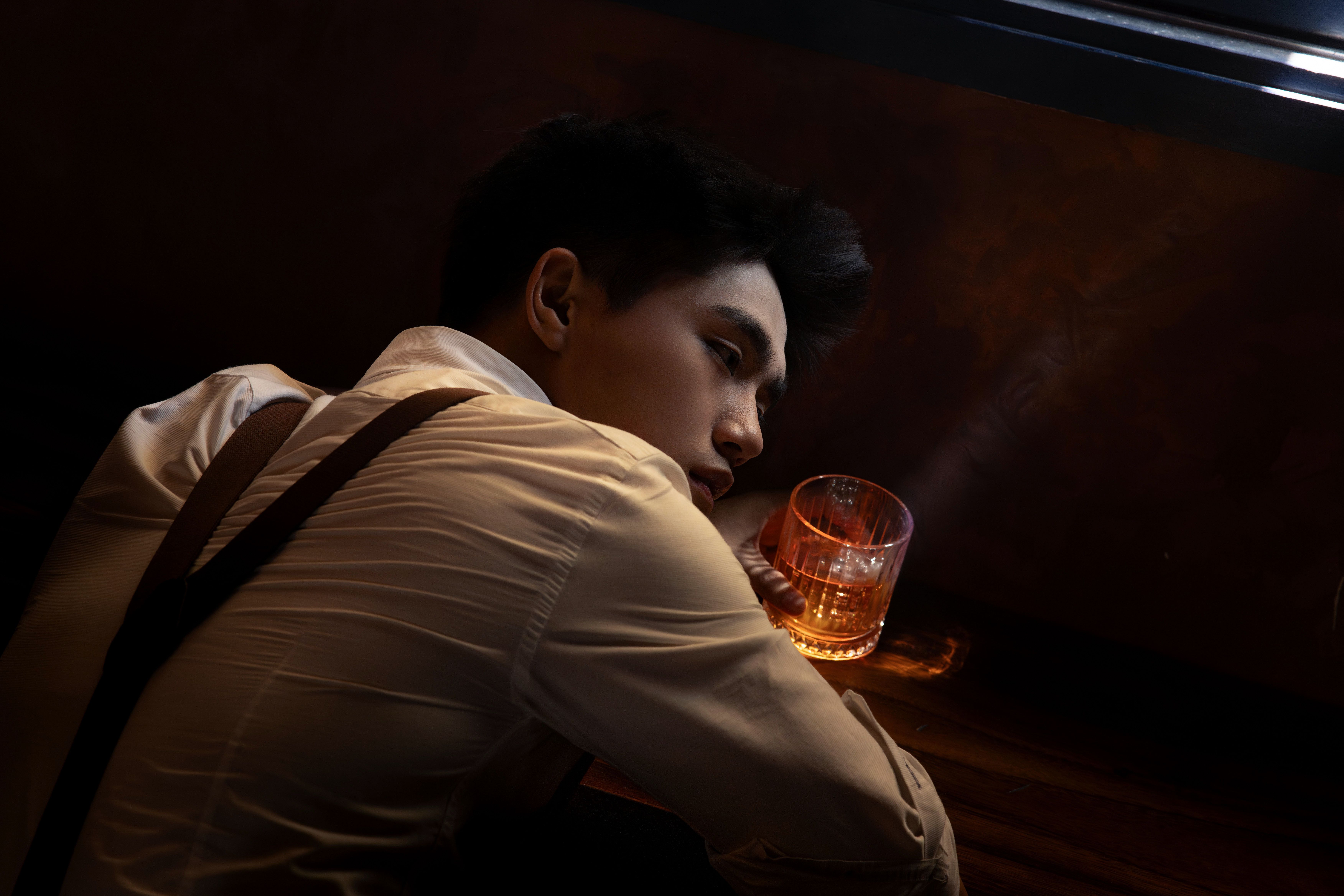 Pensive man nursing a drink alone in a shadowed bar, a quiet secretive speakeasy mood in warm low light