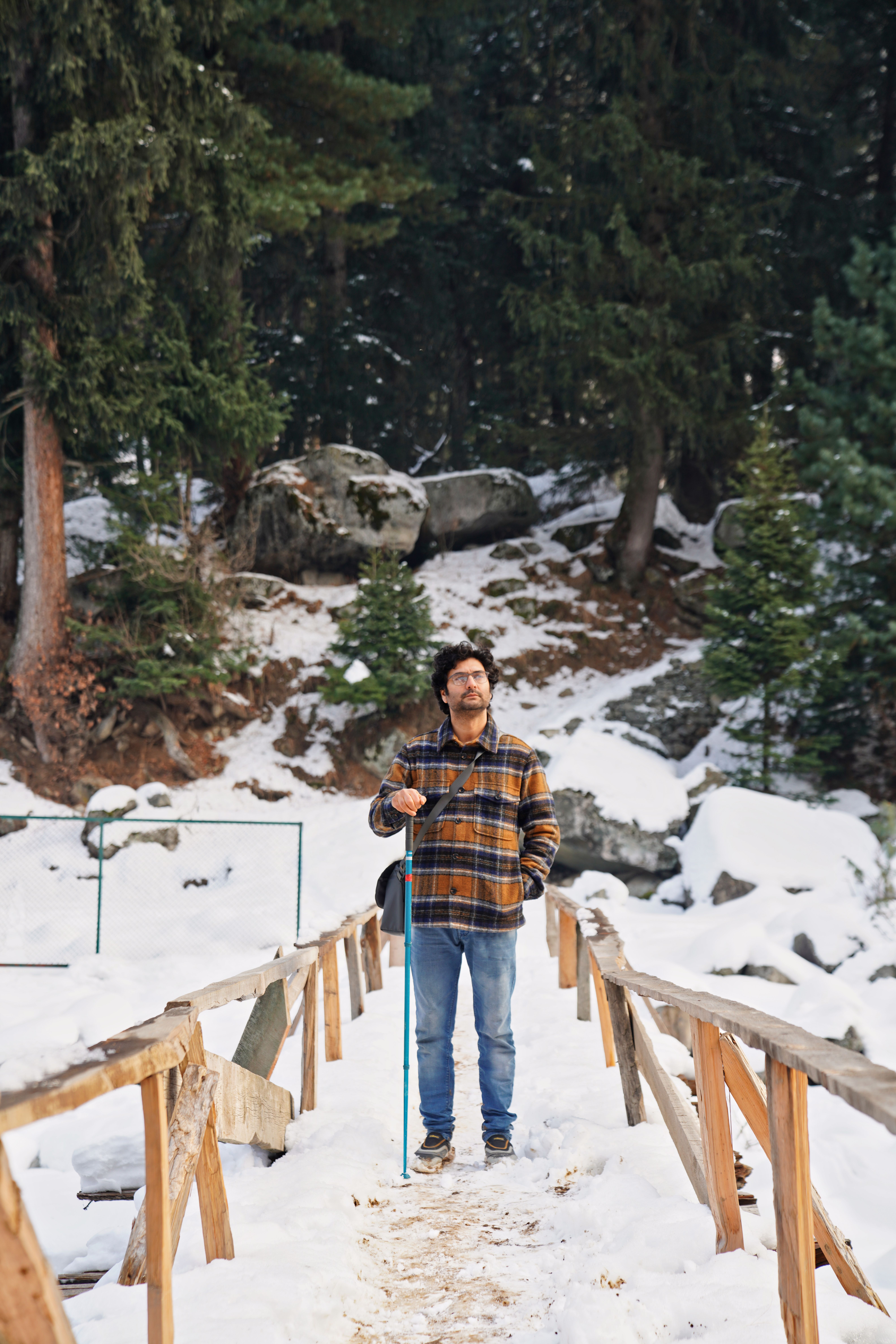 A man stands on a snowy bridge in Kashmir, surrounded by serene pine trees and winter scenery.