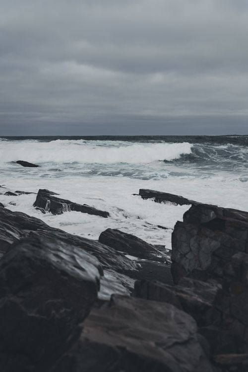 Free A moody image capturing waves crashing on a rocky shoreline under a cloudy sky. Stock Photo