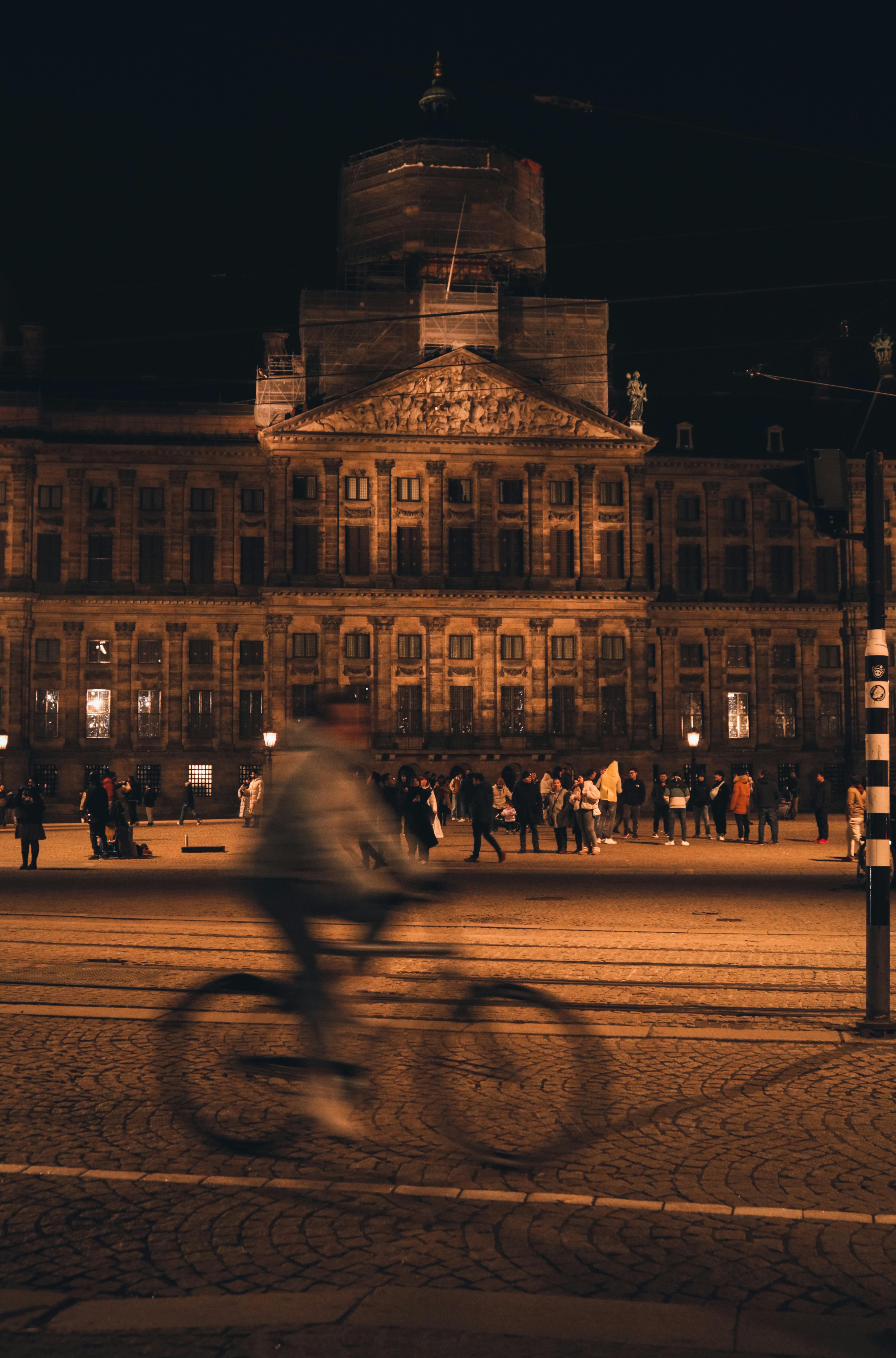 Gratis Uno scatto notturno di un ciclista che passa davanti al Palazzo Reale illuminato di Amsterdam. Foto a disposizione