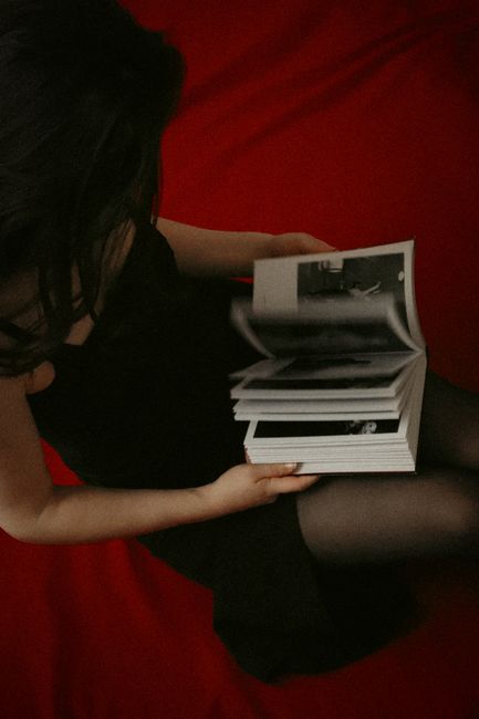 A woman in a black dress reading a book in a moody, artistic setting on a red backdrop.