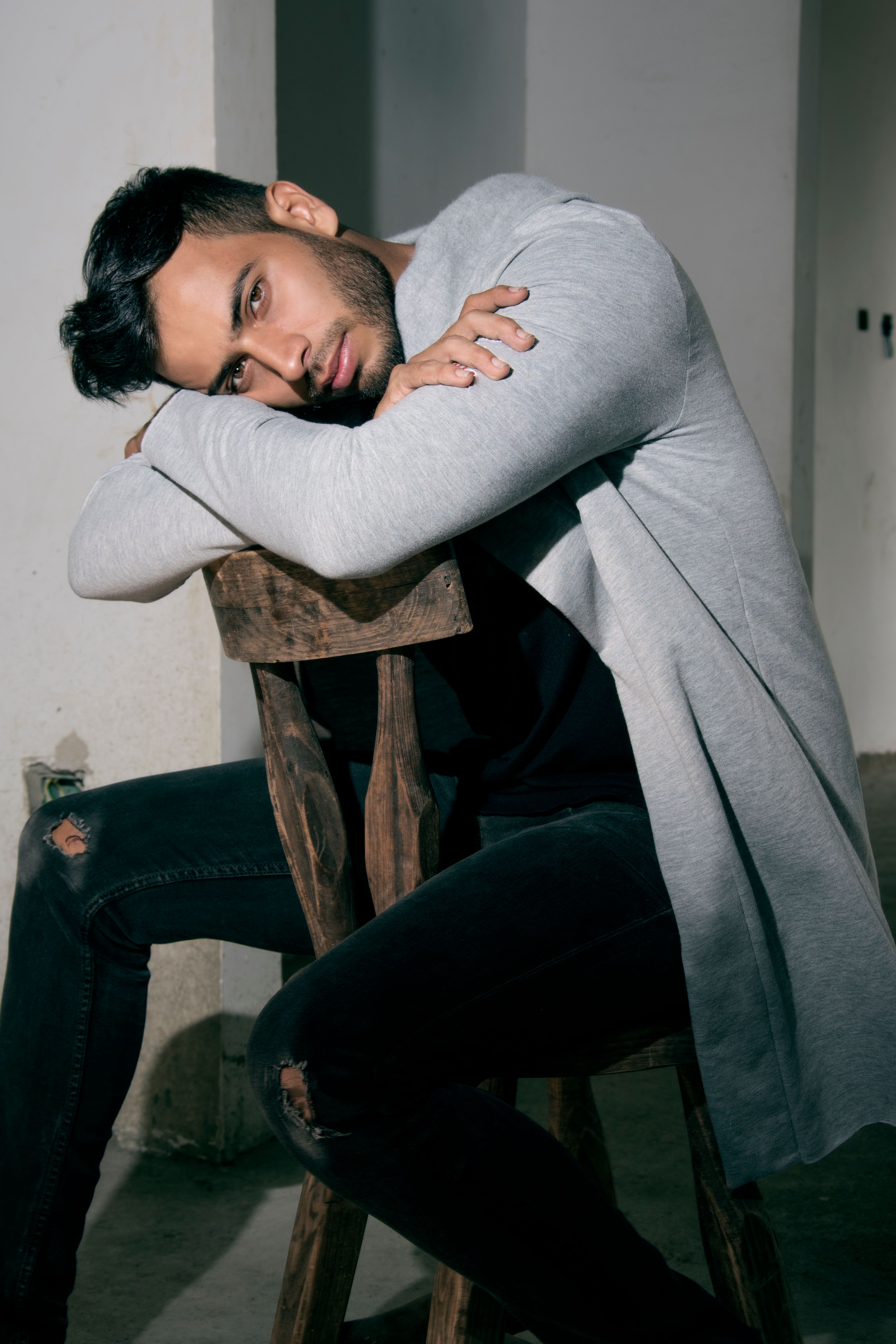 Thoughtful young man leaning on a wooden chair indoors, wearing casual attire.
