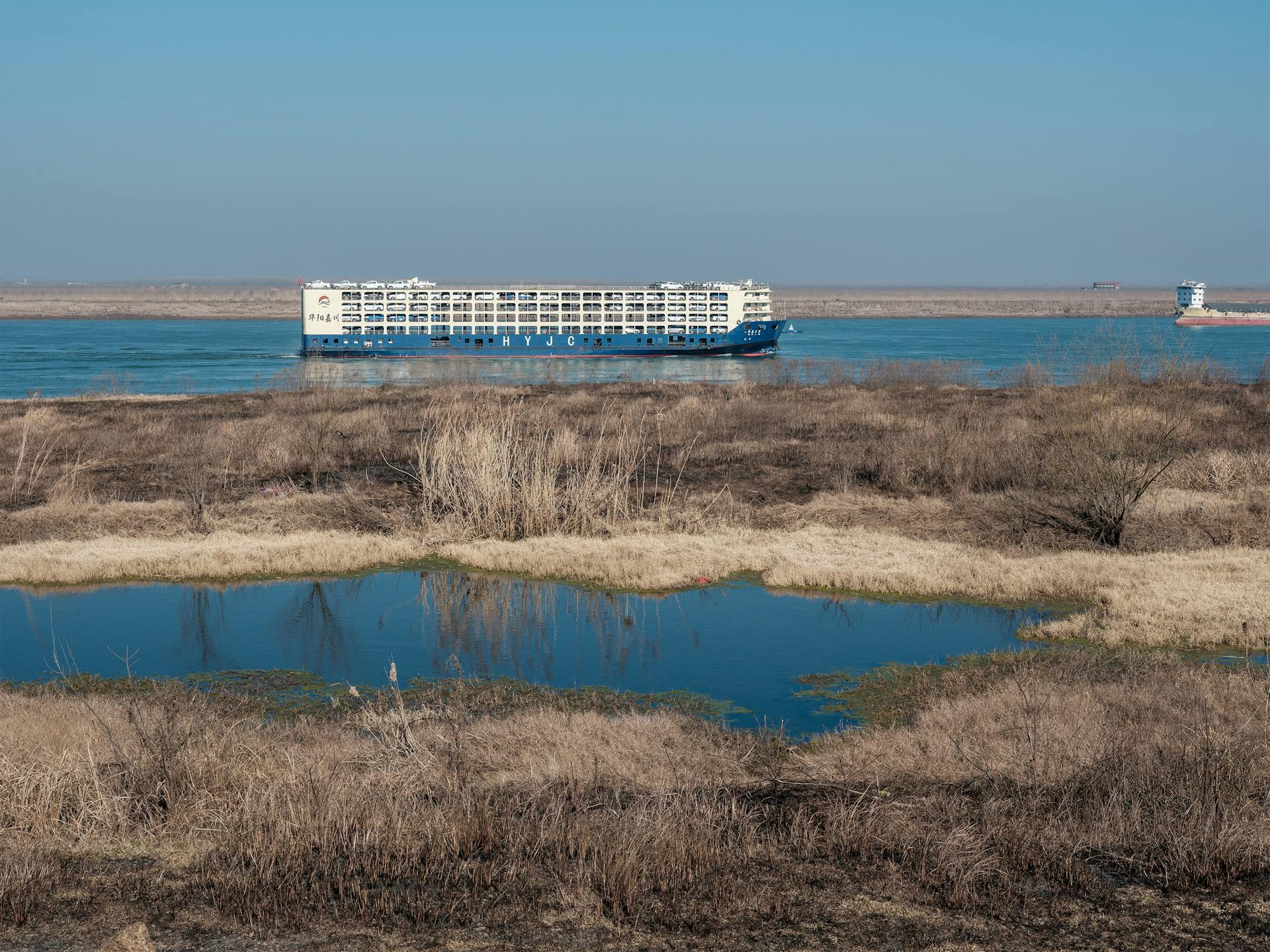 A large cargo ship navigates the serene waters of a wide river flanked by marshland.