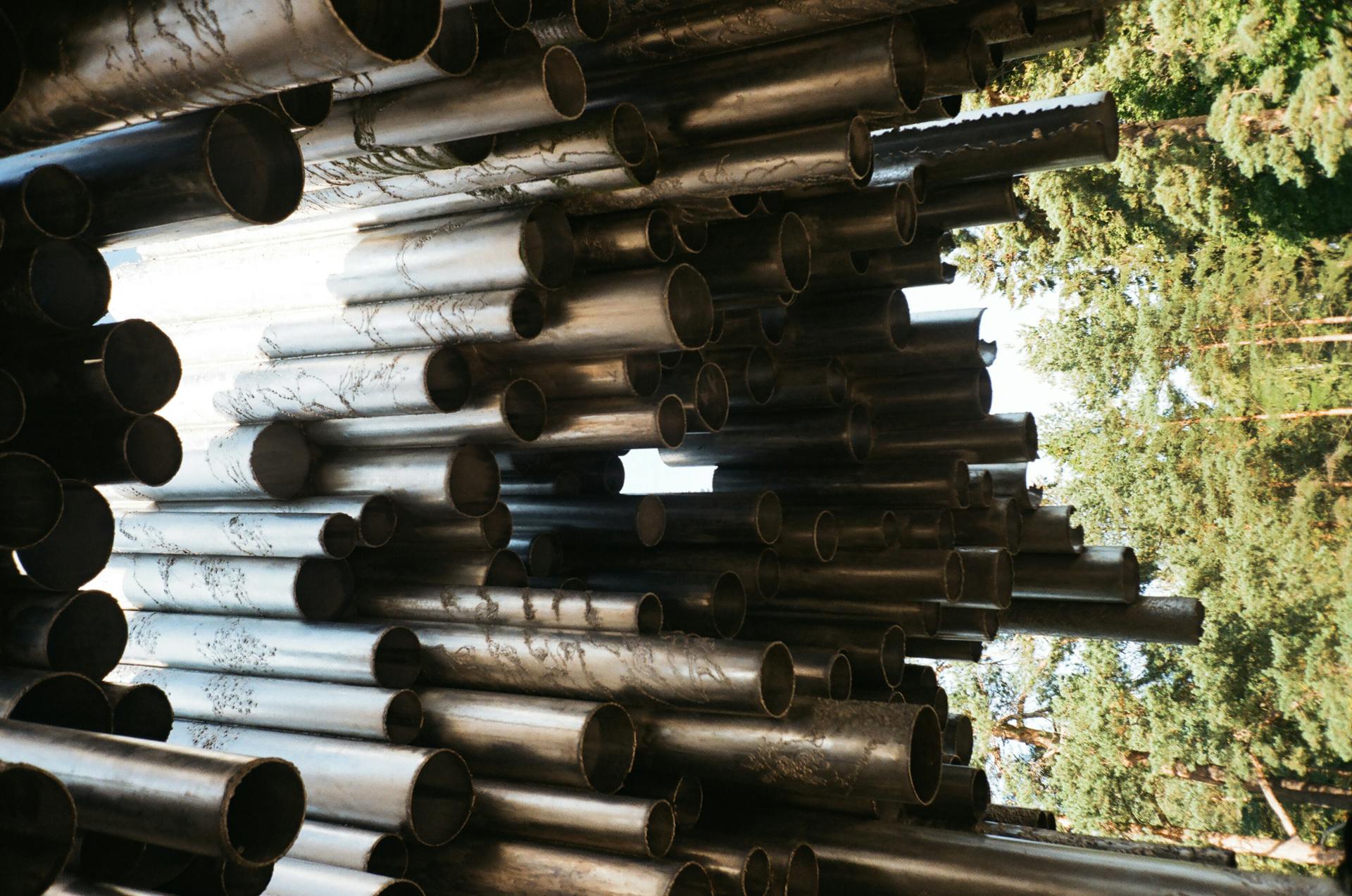 Close-up of stacked metal pipes with a background of green trees, showcasing industrial art.