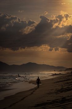 A serene beach scene with a silhouetted man walking along the shore at sunset, under dramatic clouds.