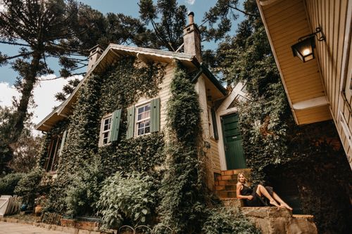 Free A woman sitting on a staircase by a charming ivy-covered cottage surrounded by greenery. Stock Photo