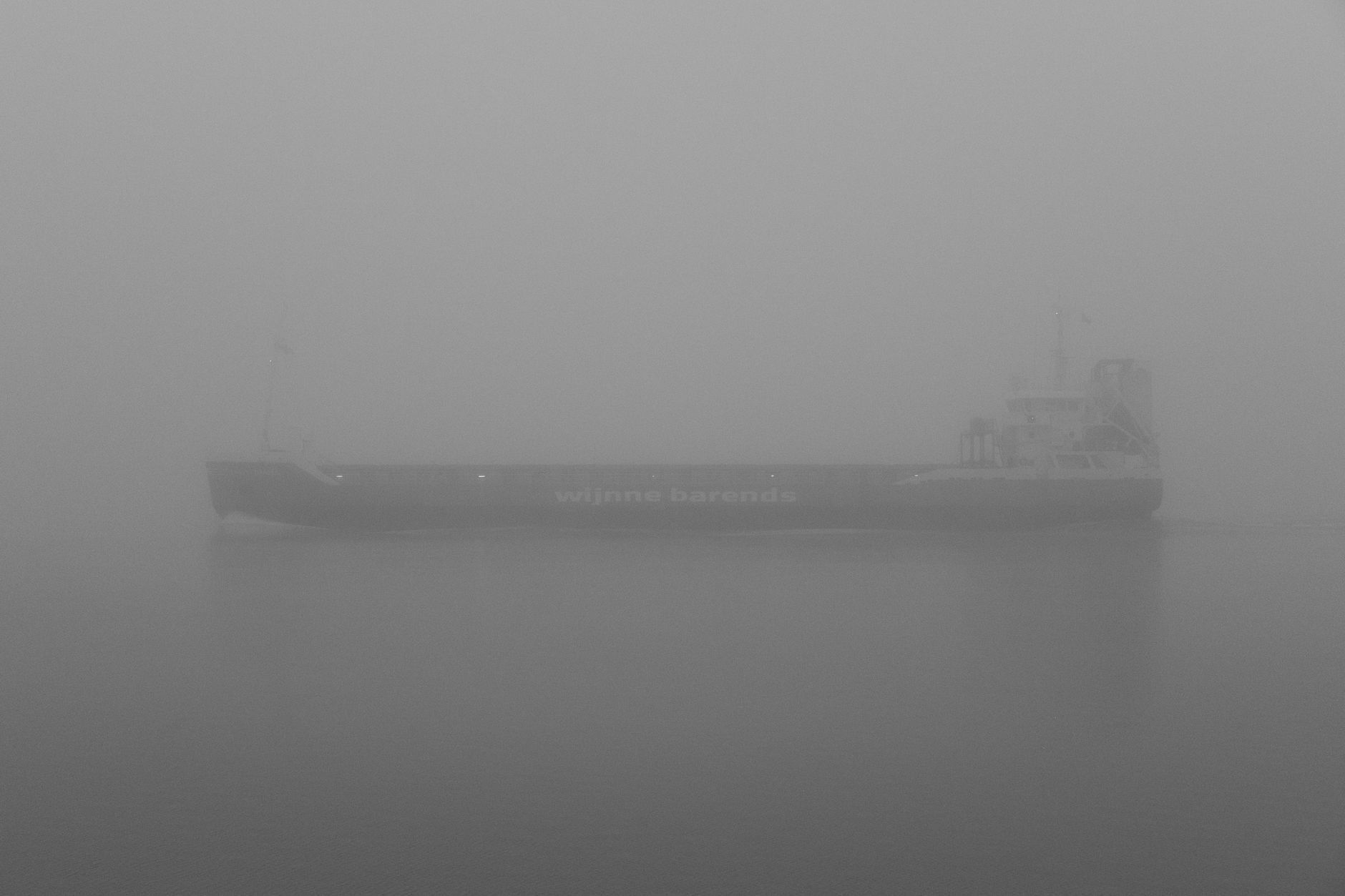 A cargo ship sailing through dense fog on calm waters, creating a serene and mysterious atmosphere.
