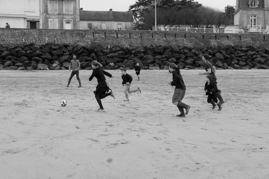 Group of children playing soccer on a sandy beach in front of a rocky seawall.