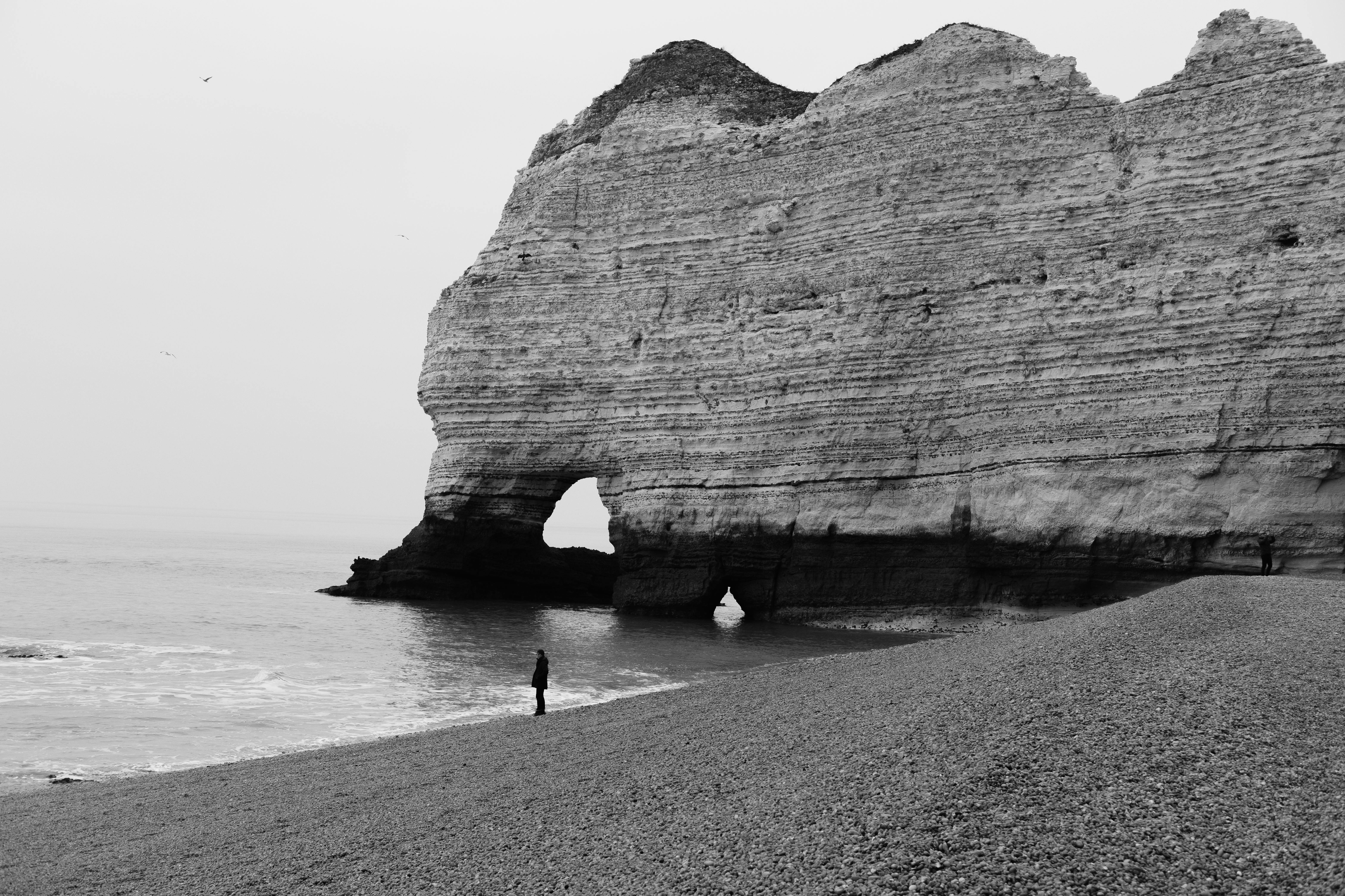 Dramatic black and white image of a person by a large sea cliff on a pebbled beach.