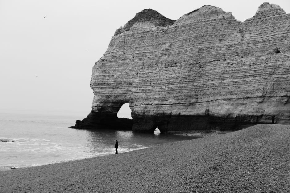 Dramatic black and white image of a person by a large sea cliff on a pebbled beach.