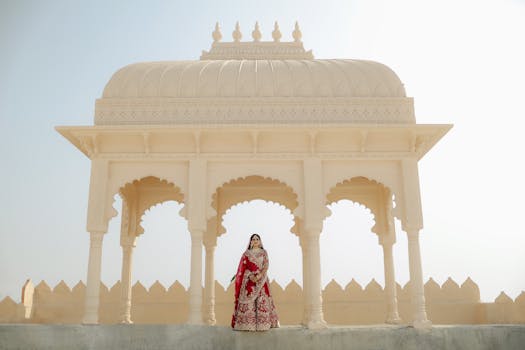 A traditional Indian bride poses elegantly at an ornate pavilion in bright daylight.
