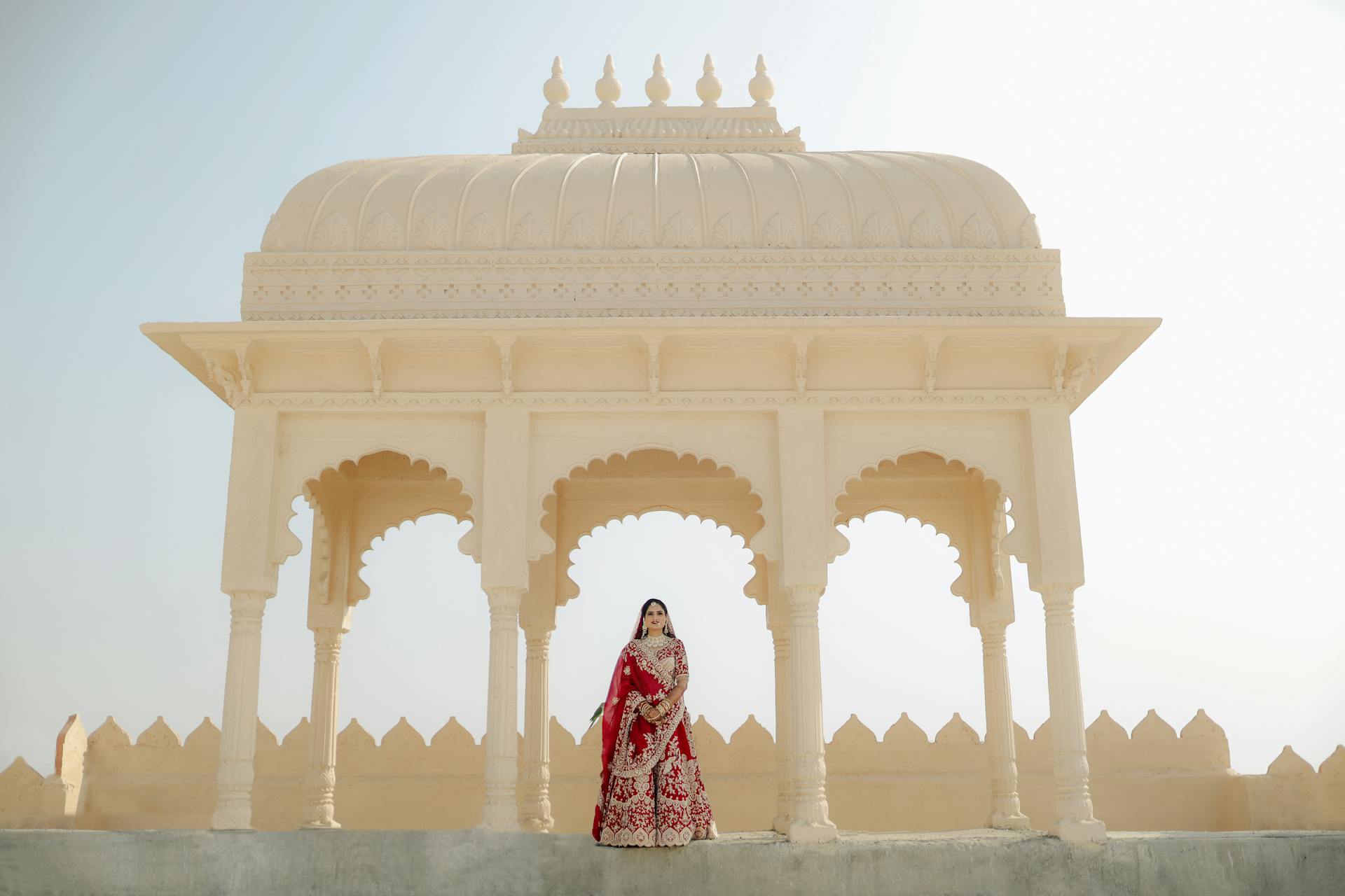 A traditional Indian bride poses elegantly at an ornate pavilion in bright daylight.