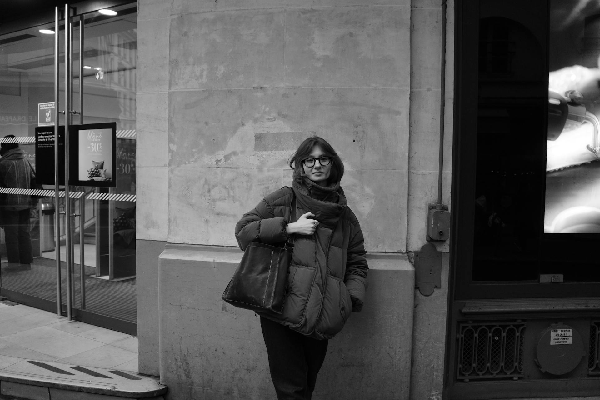 Black and white photo of a woman wearing a winter coat, standing by a storefront in a city.