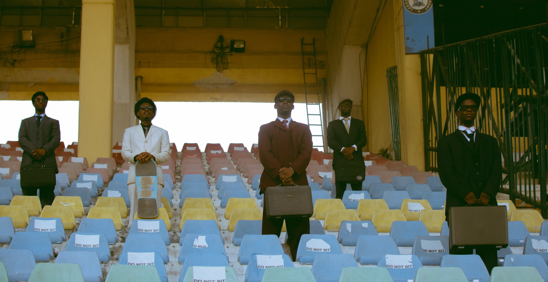 Five businessmen in suits stand in empty colorful stadium seats, holding briefcases.