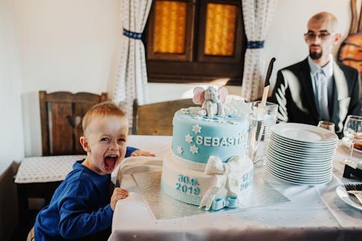 A child excitedly celebrates a birthday with a blue cake featuring an elephant topper indoors.