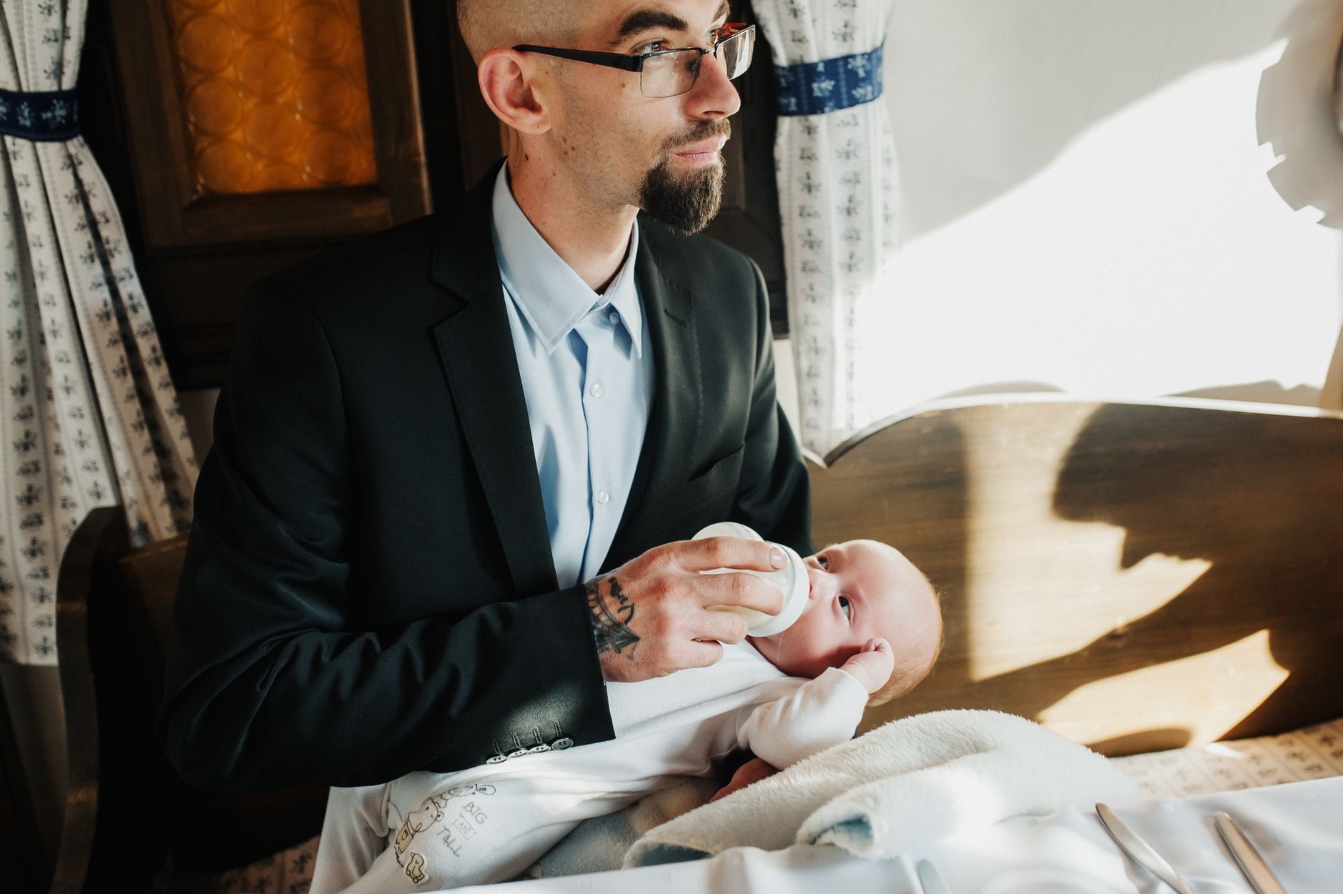 Man in suit feeding baby with bottle in cozy indoor setting, soft lighting.