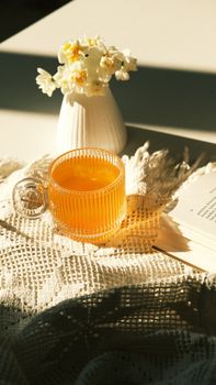 A warm cup of tea on a lace cloth with flowers and an open book in soft sunlight.