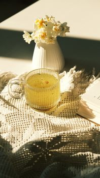 Elegant clear teacup with yellow drink beside flowers in warm morning light.