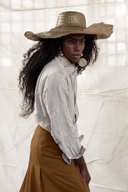 Side view of intense young African model with long hair wearing simple shirt with straw hat standing against linen backdrop