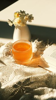 Warm morning setup with tea, a book, and flowers in sunlight.