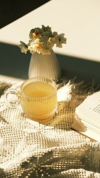 A warm morning scene featuring a cup of tea, a book, and a vase of fresh flowers.