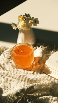 A warm sunlit setting with a glass mug of orange beverage, white flowers, and open book.