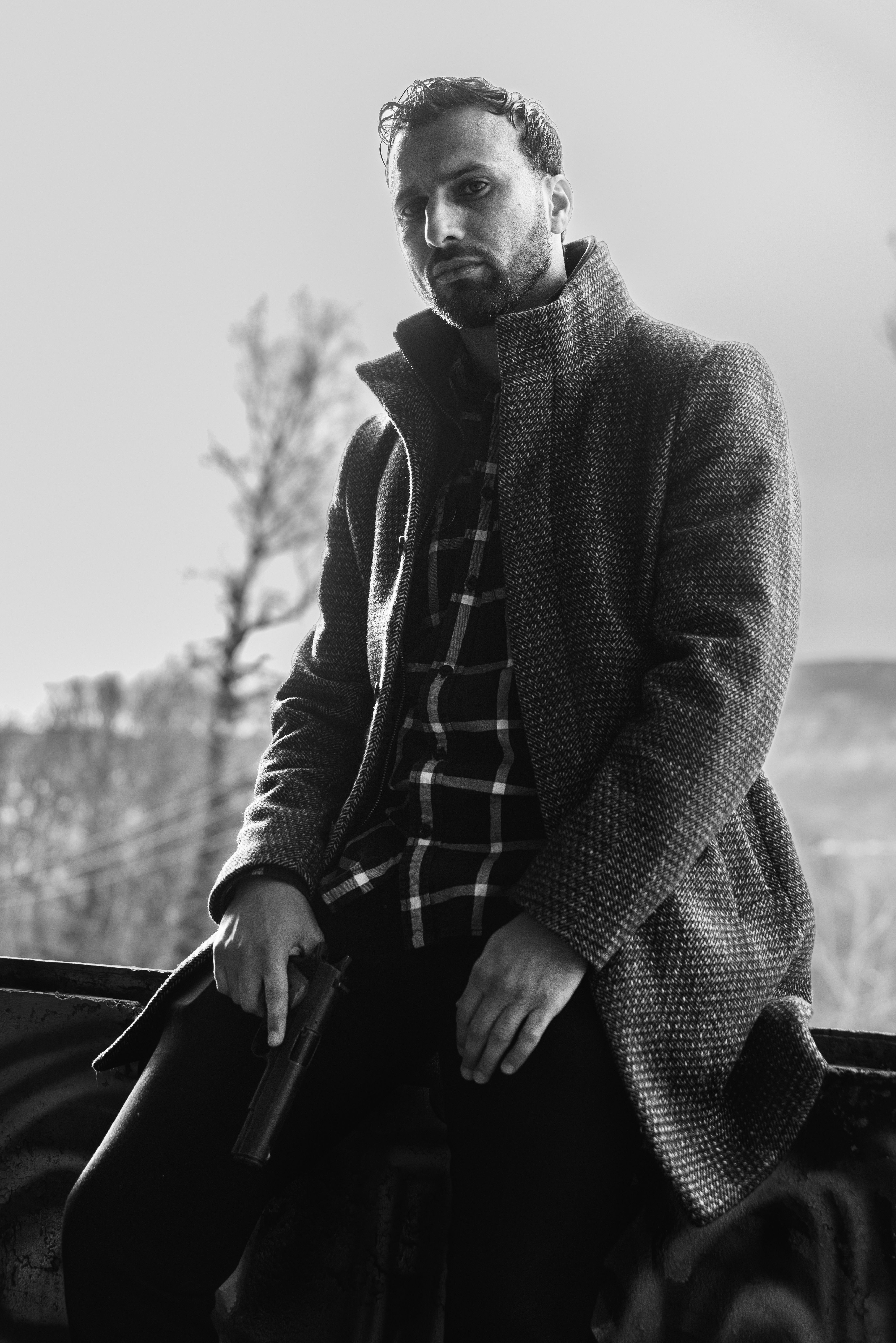 Black and white portrait of a mysterious man holding a gun in an outdoor setting.
