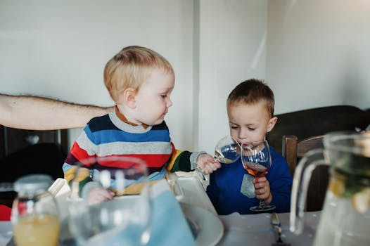 Two young children exploring beverage glasses at a family dinner table indoors.
