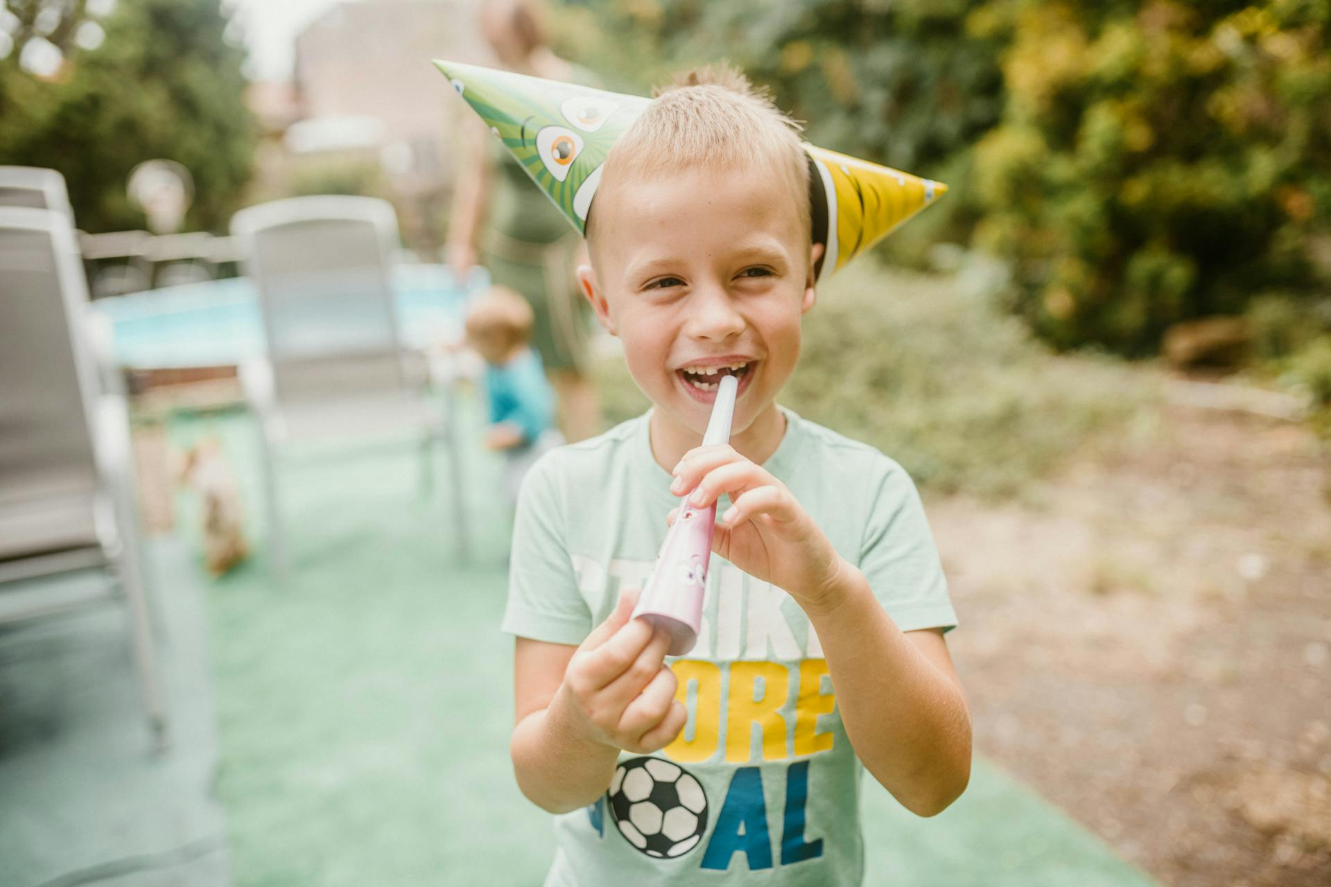 A happy child with a party hat celebrates outdoors with a noise maker.