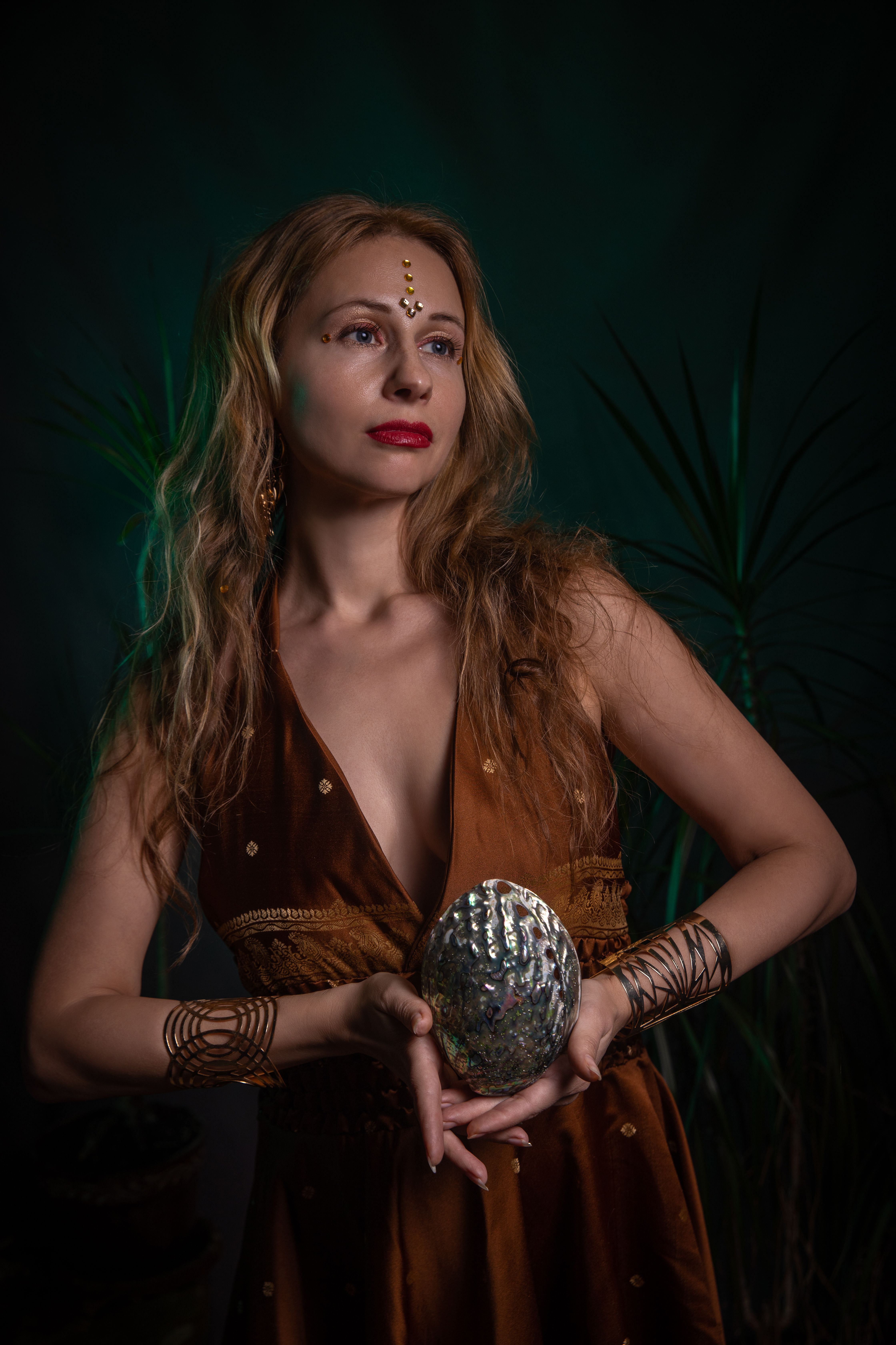 A woman in a glamorous outfit holding a decorative, reflective object indoors with a dark, moody background.