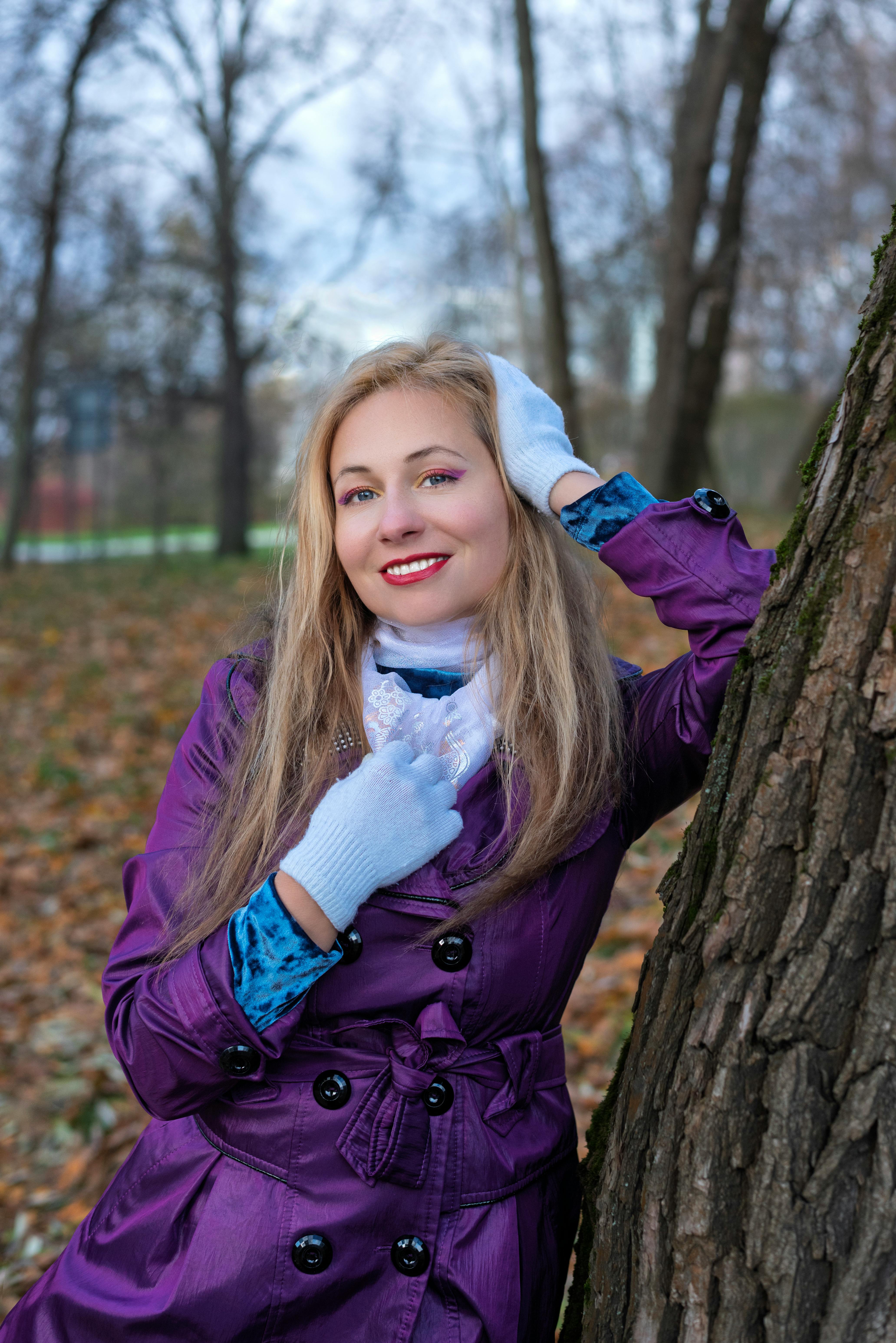 Woman in purple coat and gloves smiling in a park with autumn leaves.