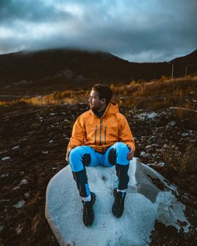 黄昏时分，一名身穿橙色夹克的男子坐在山间云雾缭绕的岩石上