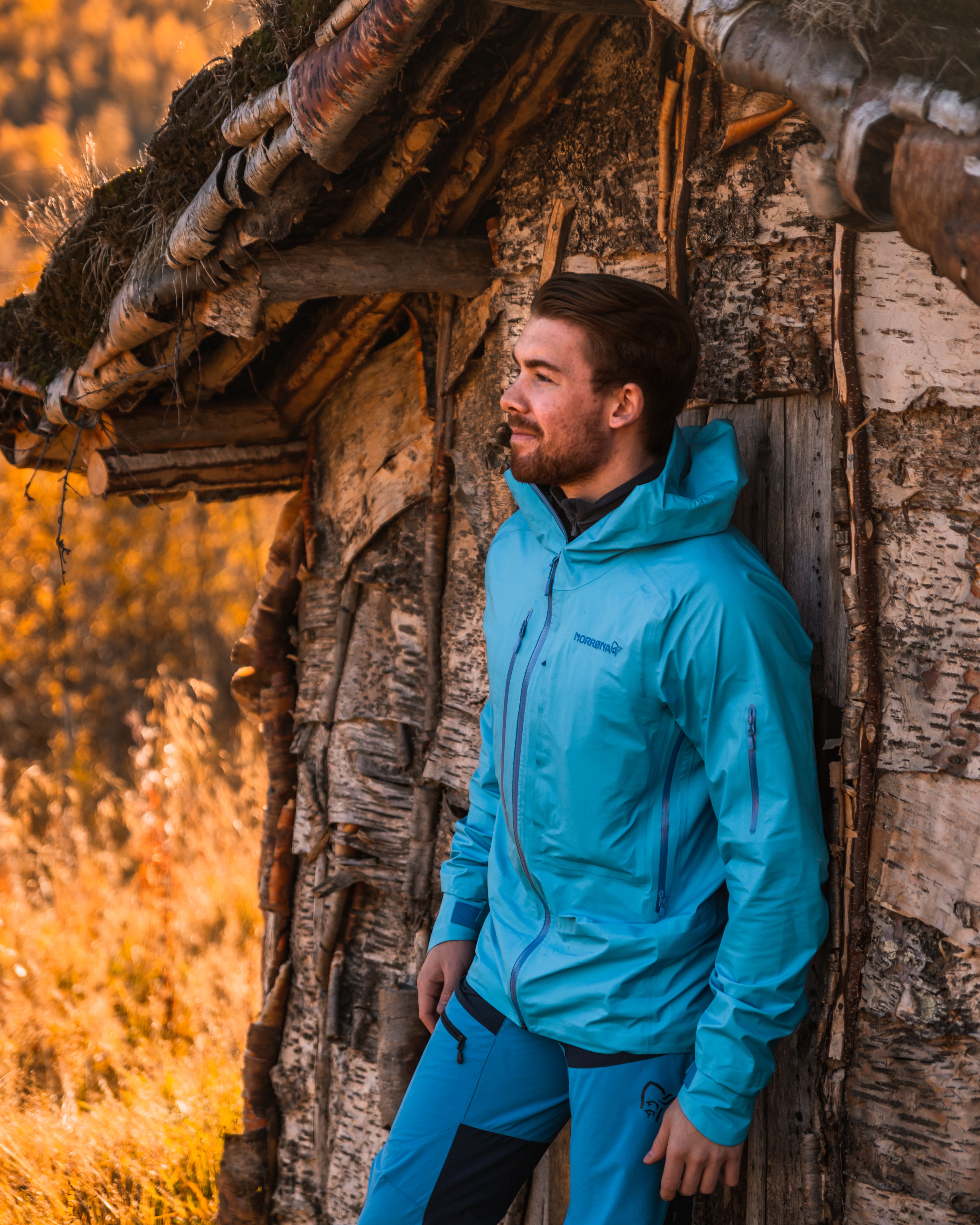 Adult man in blue jacket standing beside a rustic cabin during autumn.