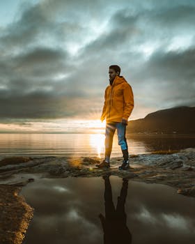 日落时分，一个穿着黄色夹克的男人站在海边，倒影映在水面上
