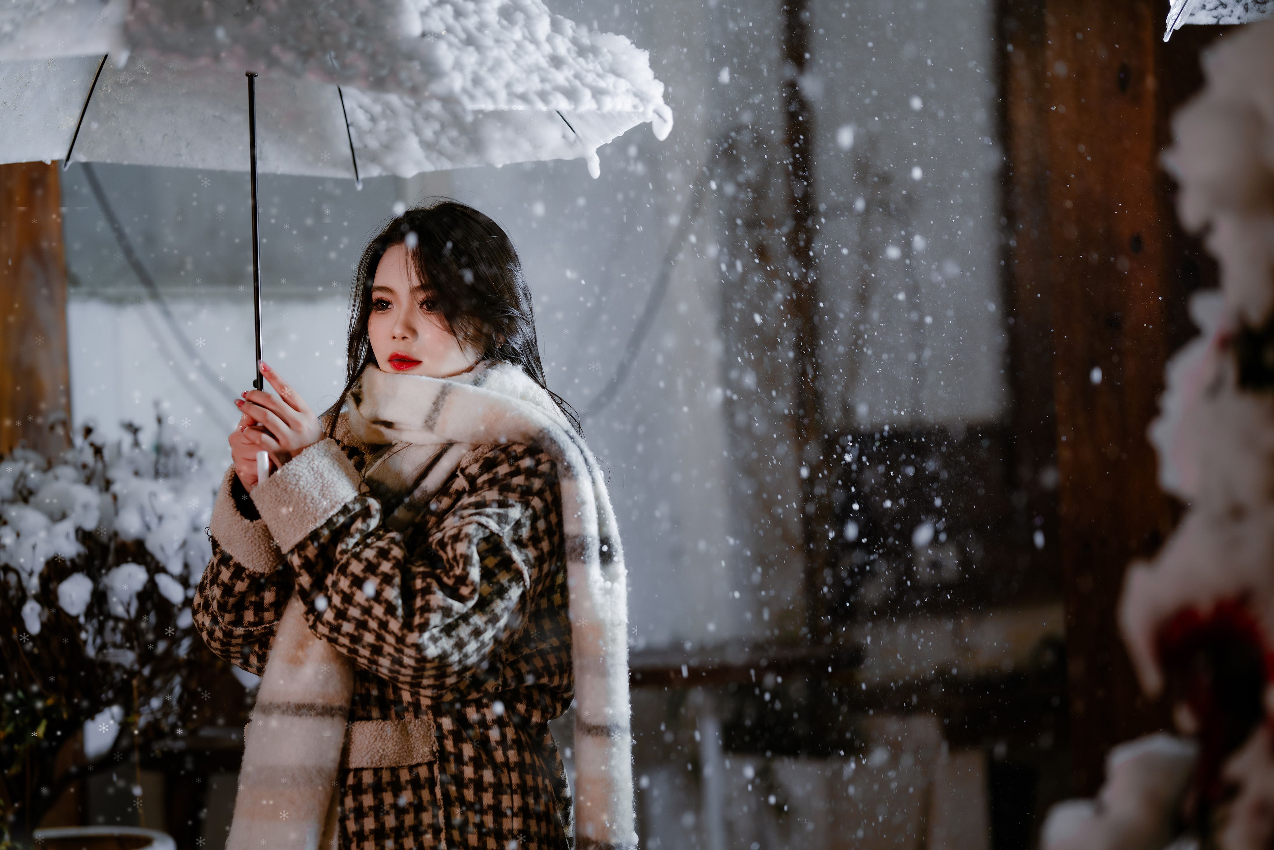 Woman standing outdoors in snowfall holding an umbrella, wrapped in warm winter clothing.