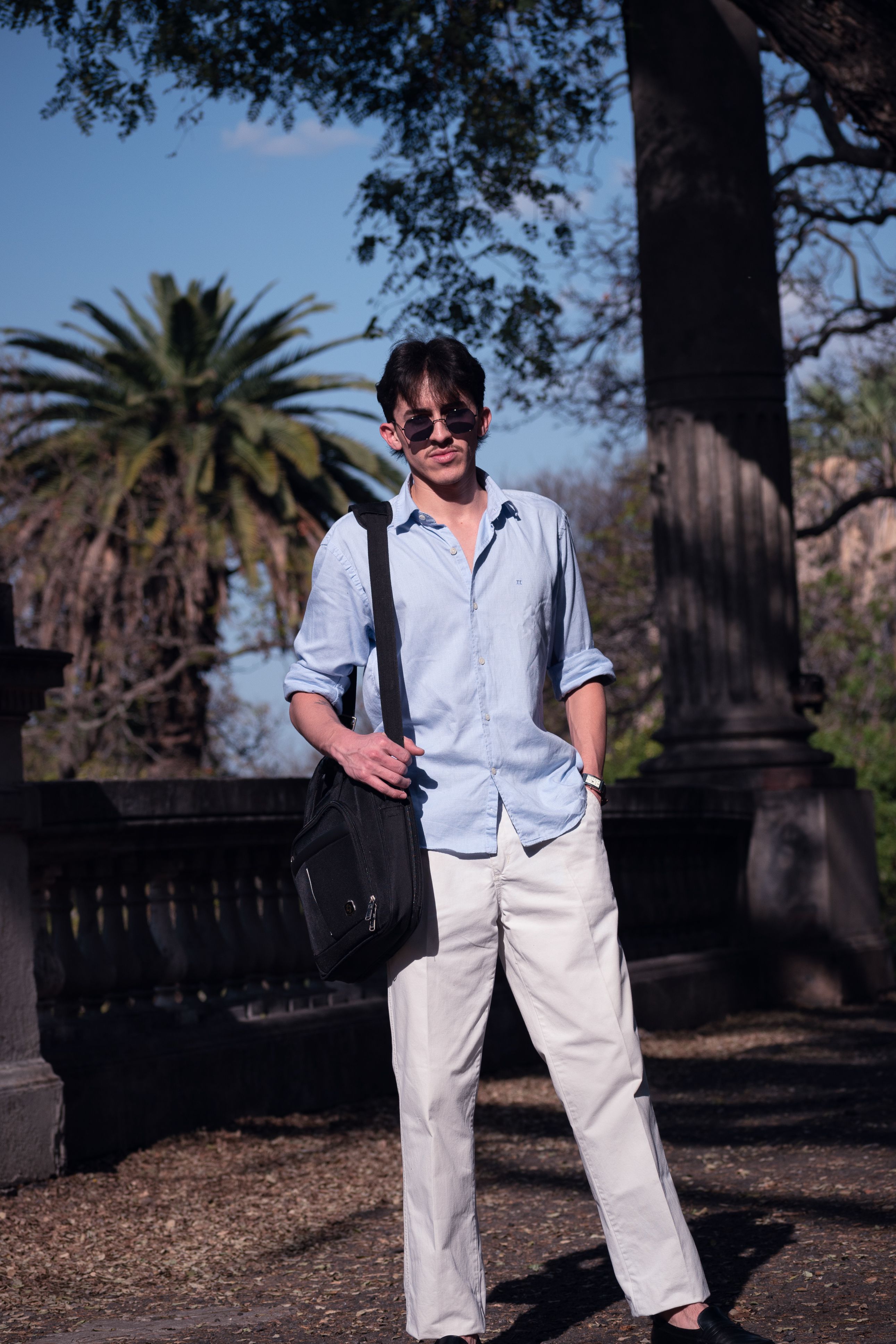 Casually dressed man poses in a sunlit Buenos Aires park with lush greenery.