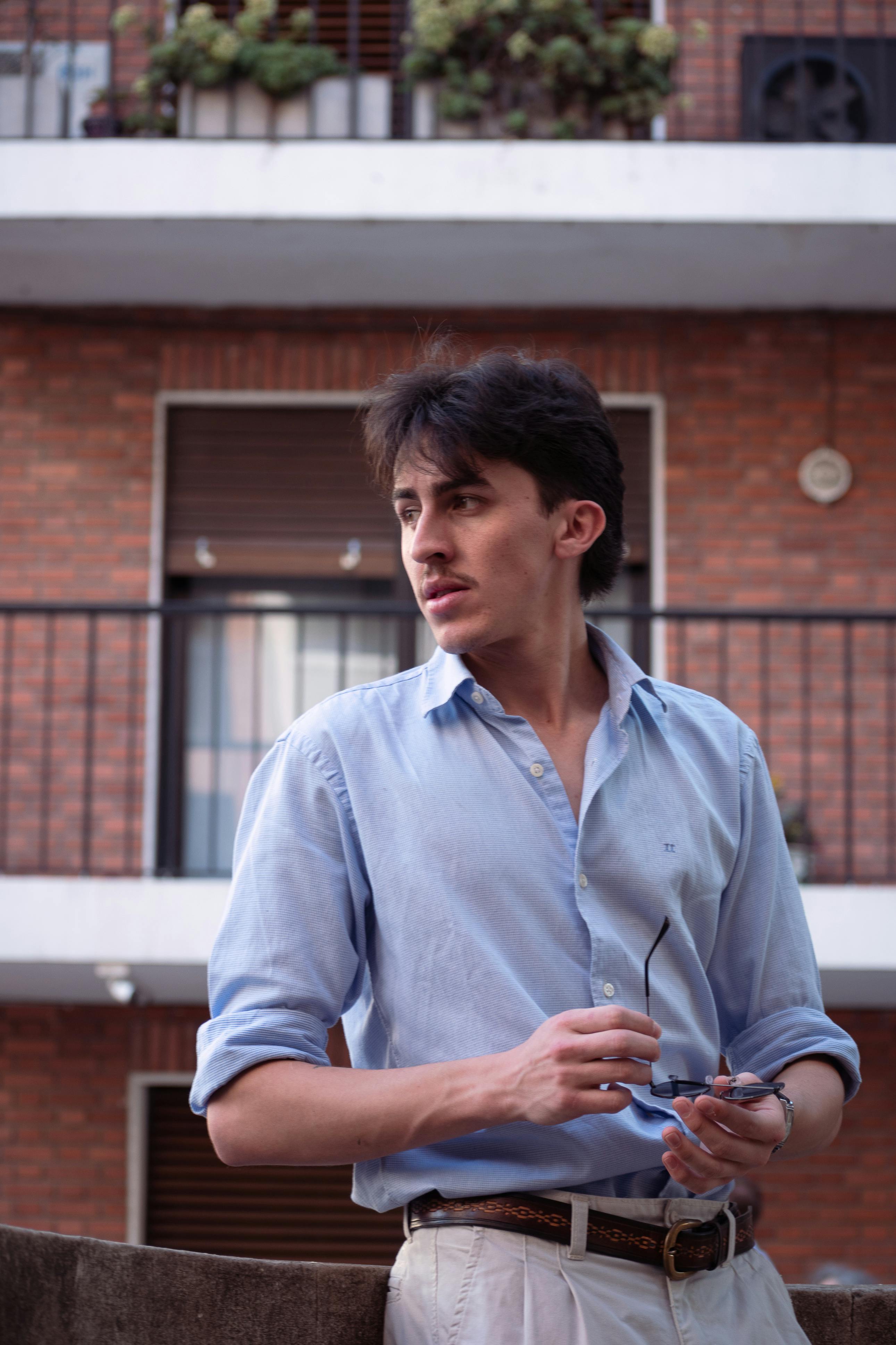 Young man in Buenos Aires stands stylishly in natural light, holding sunglasses.