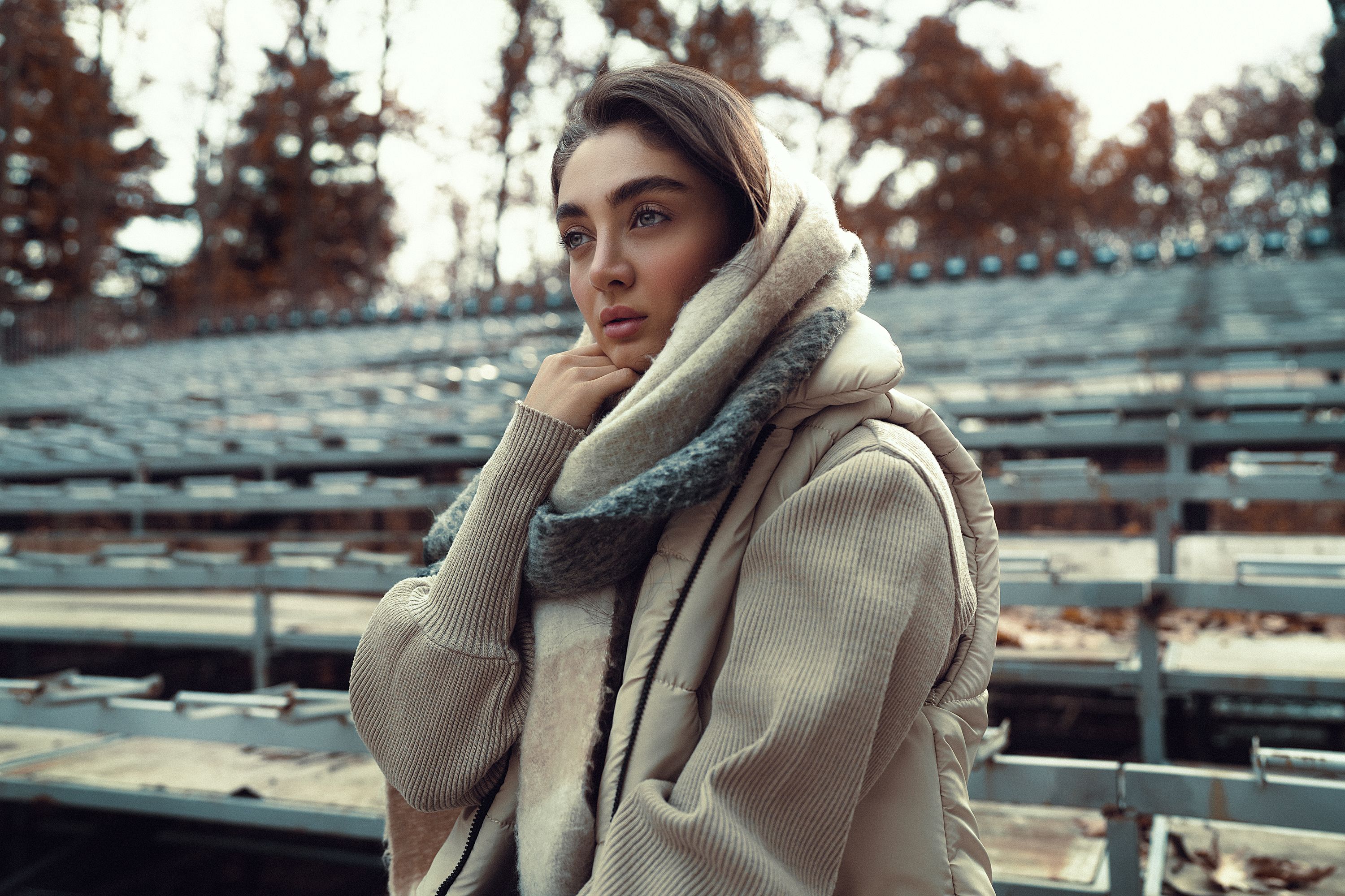 A thoughtful woman in warm attire sits outdoors during fall season.