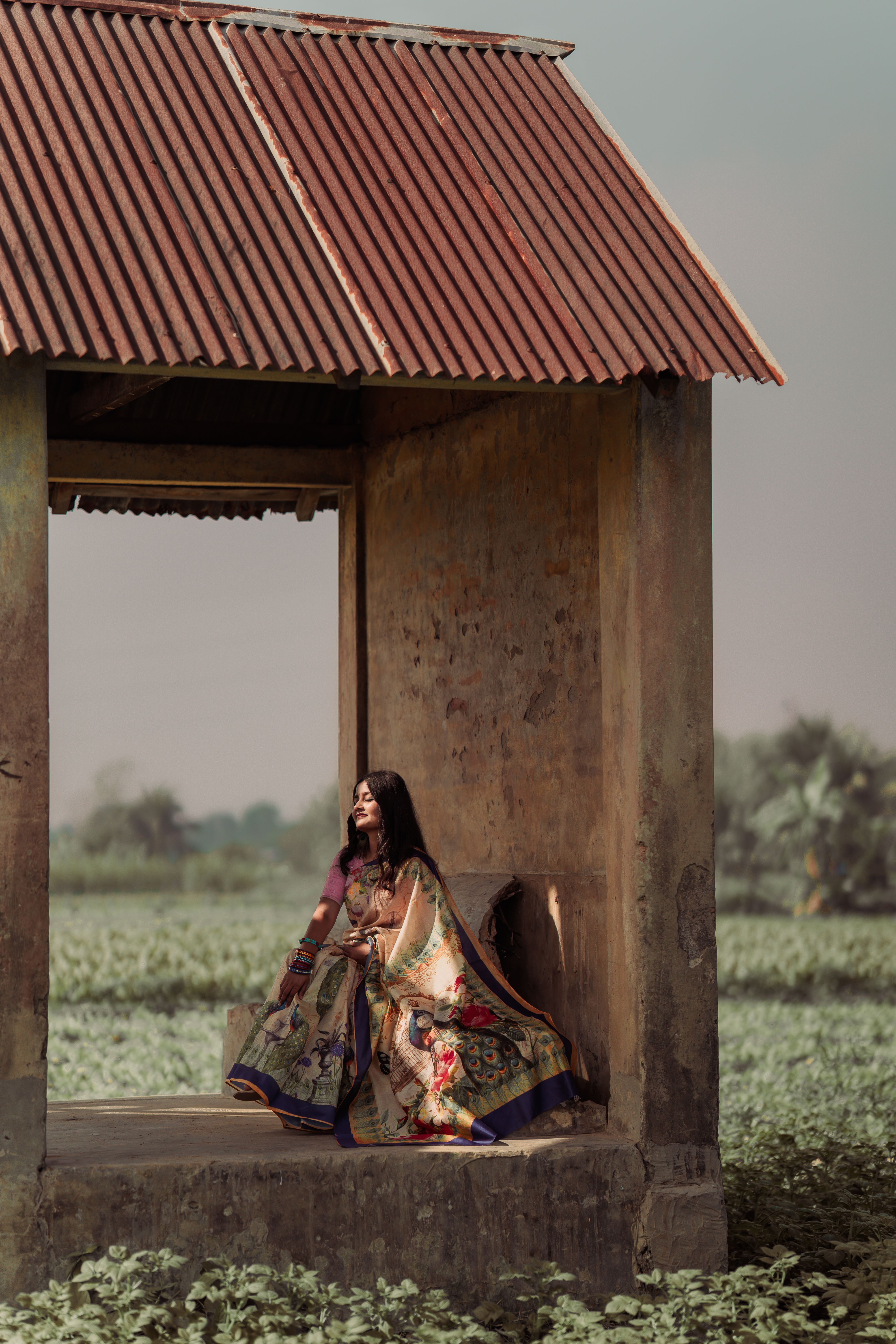 Woman in vibrant traditional dress sits in rustic structure surrounded by lush greenery.