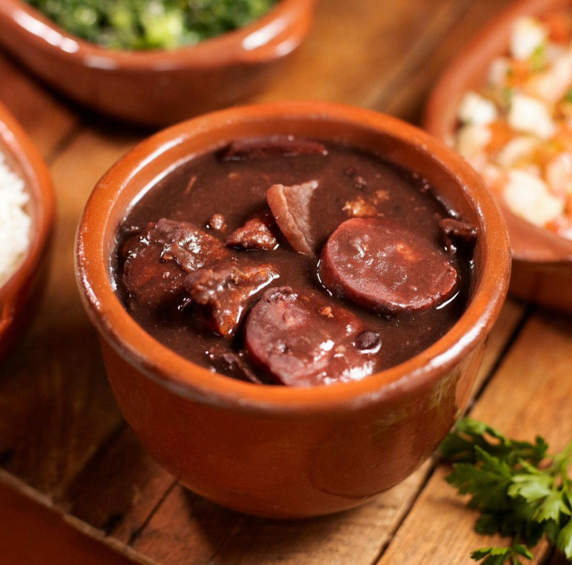 A hearty bowl of traditional Brazilian feijoada with sausages and beef in a rustic clay pot.