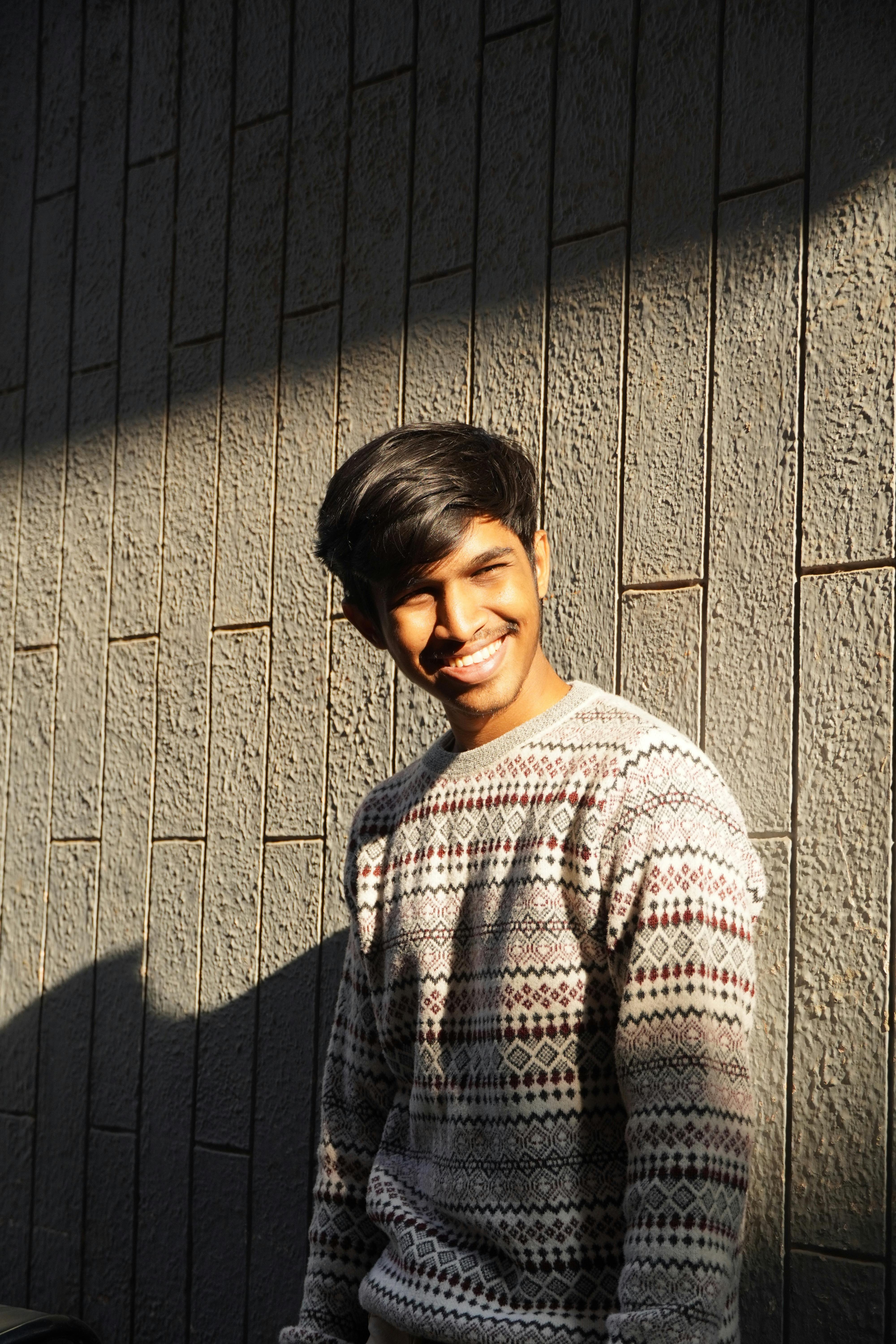 A young man smiles warmly while standing against a textured wall.
