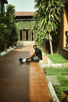 A young man relaxes in a sunlit courtyard surrounded by lush greenery in Ada Foah, Ghana.