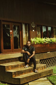 Man sitting on wooden cabin steps in Ada Foah, Ghana, with tropical plants.